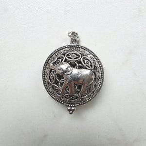 Puede incluir: Colgante plateado con un diseño de elefante dentro de un marco circular. El elefante está detallado, y el marco tiene un borde ornamentado y texturizado. El colgante incluye un lazo para colgar y un elemento decorativo inferior.