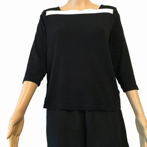 May include: Black three-quarter sleeve top with a white horizontal band across the neckline. The top is paired with black bottoms. The outfit is displayed on a mannequin, showcasing a simple, classic style.