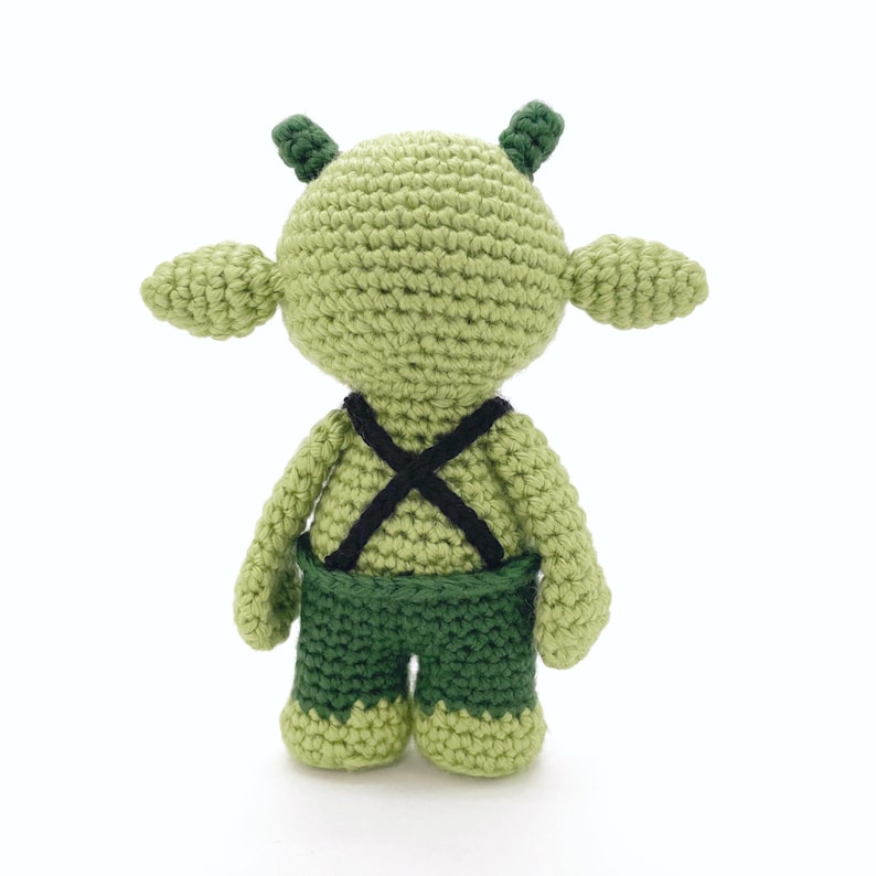 K&ouml;nnte beinhalten: Ein gr&uuml;nes H&auml;kel-Amigurumi-Spielzeug mit einem schwarzen X-f&ouml;rmigen Gurt auf dem R&uuml;cken und einer gr&uuml;nen Hose. Das Spielzeug hat gro&szlig;e Ohren und einen runden Kopf.