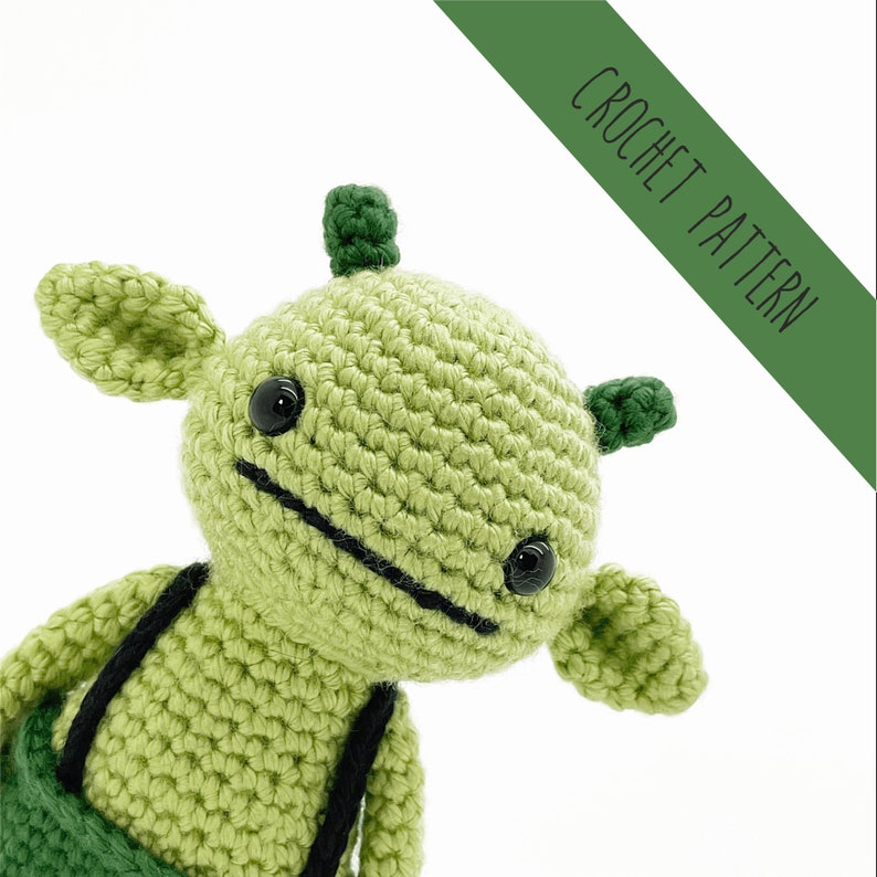 K&ouml;nnte beinhalten: Gr&uuml;nes geh&auml;keltes Amigurumi-Spielzeug mit schwarzen Augen und einem schwarzen Mund. Das Spielzeug hat gr&uuml;ne Ohren und zwei gr&uuml;ne H&ouml;rner auf dem Kopf. Das Spielzeug tr&auml;gt ein gr&uuml;nes und schwarzes Outfit.