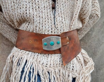 May include: A wide brown leather belt featuring a silver-toned metal buckle with three embedded turquoise stones. The belt is worn over a chunky beige knit sweater with long fringe detailing at the hem, paired with blue denim jeans.