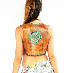 Puede incluir: Un chaleco de cuero marrón con un diseño de mandala verde y dorado en la espalda. El chaleco es corto y tiene un ribete de flecos.