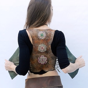 Puede incluir: Un chaleco de cuero marrón con tres diseños florales decorativos en tonos verdes, rosas y dorados. El chaleco tiene una espalda con cordones y se lleva sobre una camisa negra.