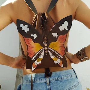 Könnte beinhalten: Ein brauner Lederrucksack mit einem Schmetterlingsdesign. Der Schmetterling hat orange, gelbe und weiße Flügel. Der Rucksack hat verstellbare Träger.