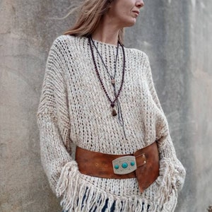 Könnte beinhalten: Eine Frau trägt einen weißen, fransigen Pullover mit einem braunen Ledergürtel mit einer silbernen Schnalle und türkisfarbenen Steinen. Sie trägt auch blaue Jeans und mehrere Halsketten.