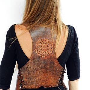 May include: A brown leather vest with a circular design on the back and a lace-up front. The vest is worn over a black shirt.