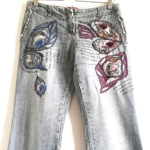 Può includere: Jeans di denim blu chiaro con ricami floreali e a forma di piume. I jeans hanno una gamba svasata e sono impreziositi da ricami colorati. I jeans sono appesi a una gruccia di legno su sfondo bianco.