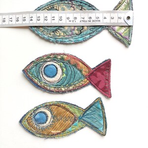 Op de afbeelding: Drie stoffen vis-applicaties met geborduurde details en gerafelde randen. Een metalen liniaal op de bovenste vis toont een lengte van ongeveer 16,5 cm. De vissen hebben diverse patronen en kleuren, waaronder blauw, bruin en rood.