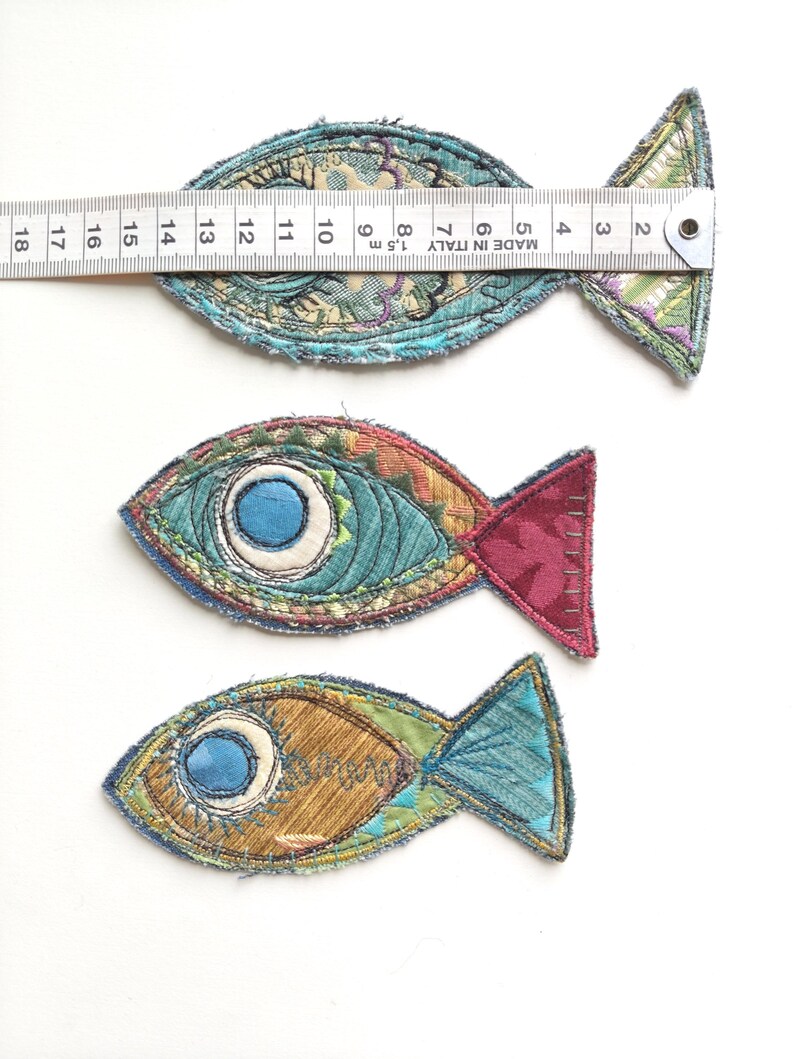 Op de afbeelding: Drie stoffen vis-applicaties met geborduurde details en gerafelde randen. Een metalen liniaal op de bovenste vis toont een lengte van ongeveer 16,5 cm. De vissen hebben diverse patronen en kleuren, waaronder blauw, bruin en rood.