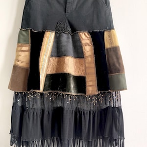 Peut inclure: Jupe noire avec un design patchwork. La partie supérieure est noire unie, passant à une section de tissus mixtes dans les tons marron, or et vert. Le bas présente un tissu noir transparent à plusieurs niveaux avec des franges.