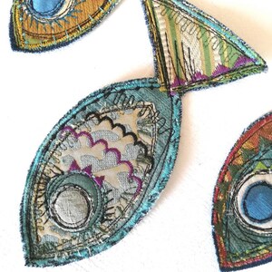 Op de afbeelding: Drie stoffen visapplicaties met ingewikkeld machinaal borduurwerk en gelaagde textielstructuren. De vissen zijn gemaakt met blauwe, groene, gouden en paarse draden, met gedetailleerde stikpatronen, geschulpte randen en cirkelvormige oogmotieven.