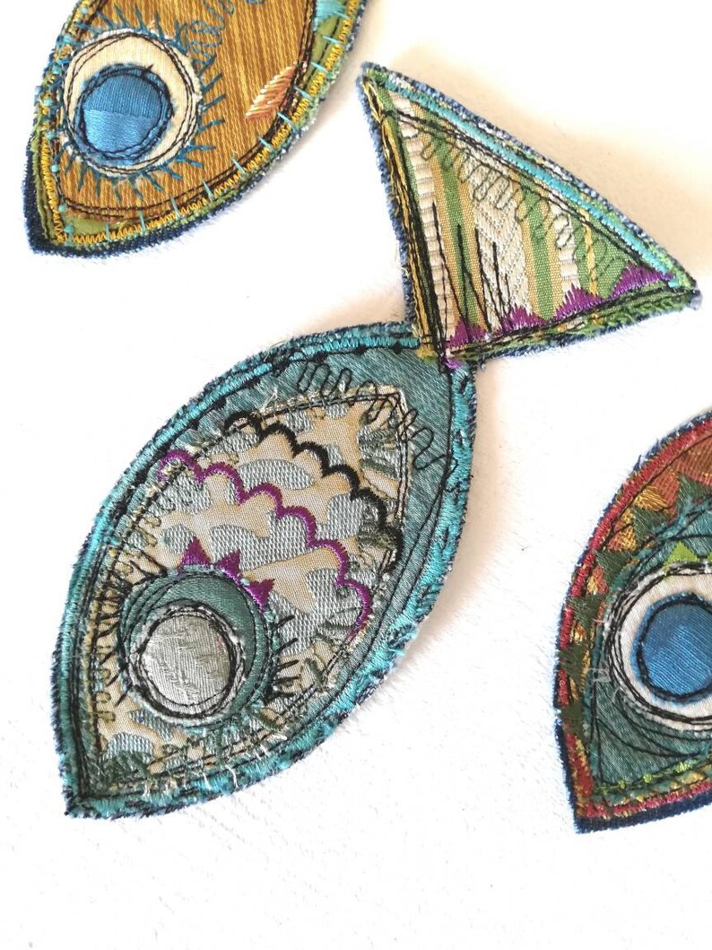 Op de afbeelding: Drie stoffen visapplicaties met ingewikkeld machinaal borduurwerk en gelaagde textielstructuren. De vissen zijn gemaakt met blauwe, groene, gouden en paarse draden, met gedetailleerde stikpatronen, geschulpte randen en cirkelvormige oogmotieven.