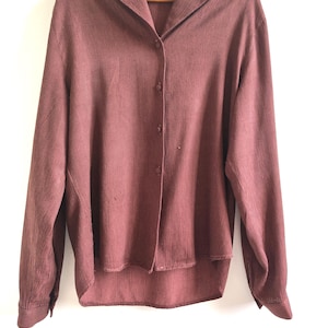 Può includere: Una camicia a maniche lunghe di colore malva tenue, con colletto classico e chiusura con bottoni. La camicia è realizzata in un tessuto strutturato ed è appesa a una gruccia di legno. Lo sfondo è bianco. La camicia ha una vestibilità rilassata.