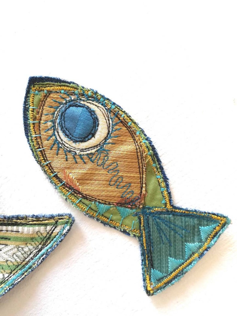 Op de afbeelding: Een stoffen vis-applicatie met een goudbruin lichaam, een groot blauw cirkelvormig oog en accenten van blauwgroen en geel borduurwerk. De staart is van donkerblauwgroene stof met geometrisch borduurwerk en een gele gestikte rand.