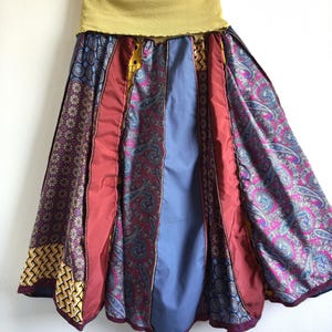 Può includere: Gonna patchwork con fascia in vita color senape e silhouette svasata. La gonna presenta pannelli di vari tessuti fantasia nei toni del rosso, blu, viola e giallo. La gonna è appesa a una gruccia su sfondo bianco.