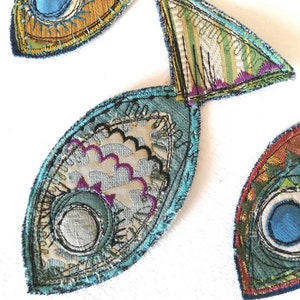 Op de afbeelding: Drie stoffen visapplicaties met ingewikkeld machinaal borduurwerk en gelaagde textielstructuren. De vissen zijn gemaakt met blauwe, groene, gouden en paarse draden, met gedetailleerde stikpatronen, geschulpte randen en cirkelvormige oogmotieven.