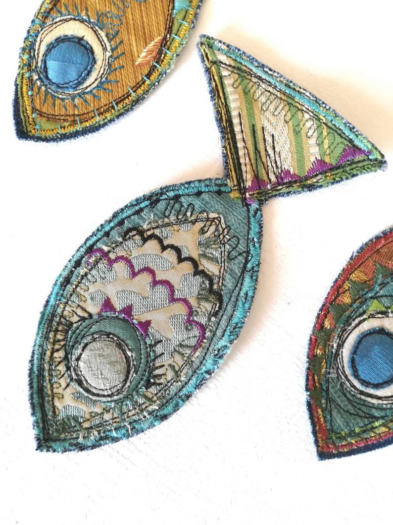 Op de afbeelding: Drie stoffen visapplicaties met ingewikkeld machinaal borduurwerk en gelaagde textielstructuren. De vissen zijn gemaakt met blauwe, groene, gouden en paarse draden, met gedetailleerde stikpatronen, geschulpte randen en cirkelvormige oogmotieven.