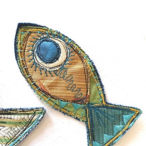 Op de afbeelding: Een stoffen vis-applicatie met een goudbruin lichaam, een groot blauw cirkelvormig oog en accenten van blauwgroen en geel borduurwerk. De staart is van donkerblauwgroene stof met geometrisch borduurwerk en een gele gestikte rand.