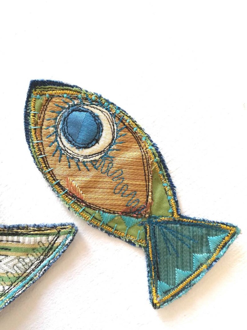 Op de afbeelding: Een stoffen vis-applicatie met een goudbruin lichaam, een groot blauw cirkelvormig oog en accenten van blauwgroen en geel borduurwerk. De staart is van donkerblauwgroene stof met geometrisch borduurwerk en een gele gestikte rand.