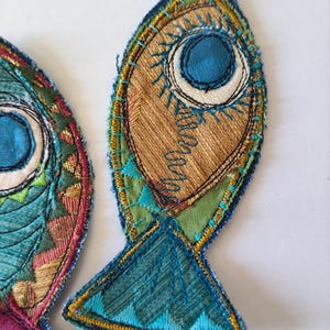 Op de afbeelding: Twee stoffen visapplicaties met ingewikkeld borduurwerk en patchwork. De vissen zijn gemaakt van blauwgroene, groene, gouden en bruine stoffen, geaccentueerd met decoratieve stiksels, zichtbare draadstructuren en een opvallend rond oogontwerp.