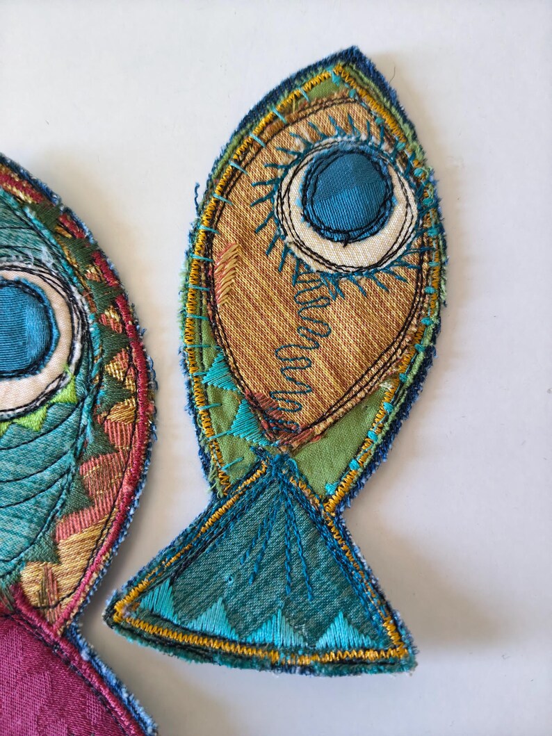 Op de afbeelding: Twee stoffen visapplicaties met ingewikkeld borduurwerk en patchwork. De vissen zijn gemaakt van blauwgroene, groene, gouden en bruine stoffen, geaccentueerd met decoratieve stiksels, zichtbare draadstructuren en een opvallend rond oogontwerp.