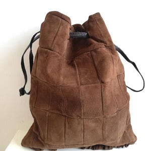 May include: A brown suede drawstring bag with a patchwork design. The bag has a rounded shape and a black drawstring closure. The suede has a soft, textured appearance. The bag is designed to be carried over the shoulder.