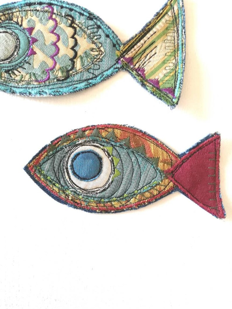 Op de afbeelding: Twee stoffen visapplicaties met ingewikkeld borduurwerk en gemengde textielpatronen. De bovenste vis heeft lichtblauwe, cr&egrave;mekleurige en paarse tinten, terwijl de onderste vis een opvallend blauw oog, een groenblauw lichaam en een donkerrode staart heeft.
