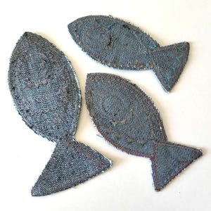 Op de afbeelding: Drie visvormige stoffen patches gemaakt van blauw denim materiaal. Elke patch is voorzien van decoratieve stiksels die het vissenlichaam, een cirkelvormig oog en een staartvin omlijnen. De randen zijn afgewerkt met een donkere draad.