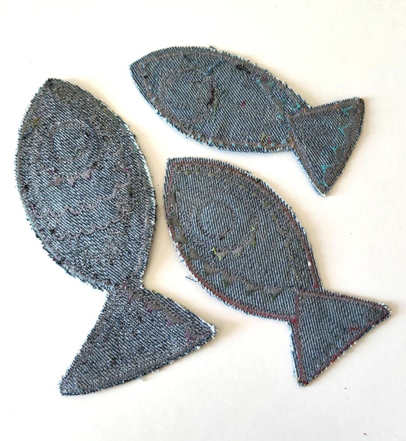 Op de afbeelding: Drie visvormige stoffen patches gemaakt van blauw denim materiaal. Elke patch is voorzien van decoratieve stiksels die het vissenlichaam, een cirkelvormig oog en een staartvin omlijnen. De randen zijn afgewerkt met een donkere draad.