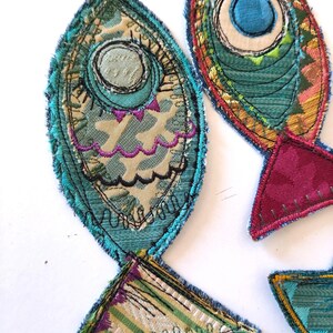 Op de afbeelding: Twee stoffen visapplicaties gemaakt van gelaagde textielresten in tinten blauwgroen, magenta en cr&egrave;me. De visvormen zijn omlijnd met donkerblauw stiksel en bevatten cirkelvormige oogdetails, decoratief schulprandborduurwerk en rafelige randen.
