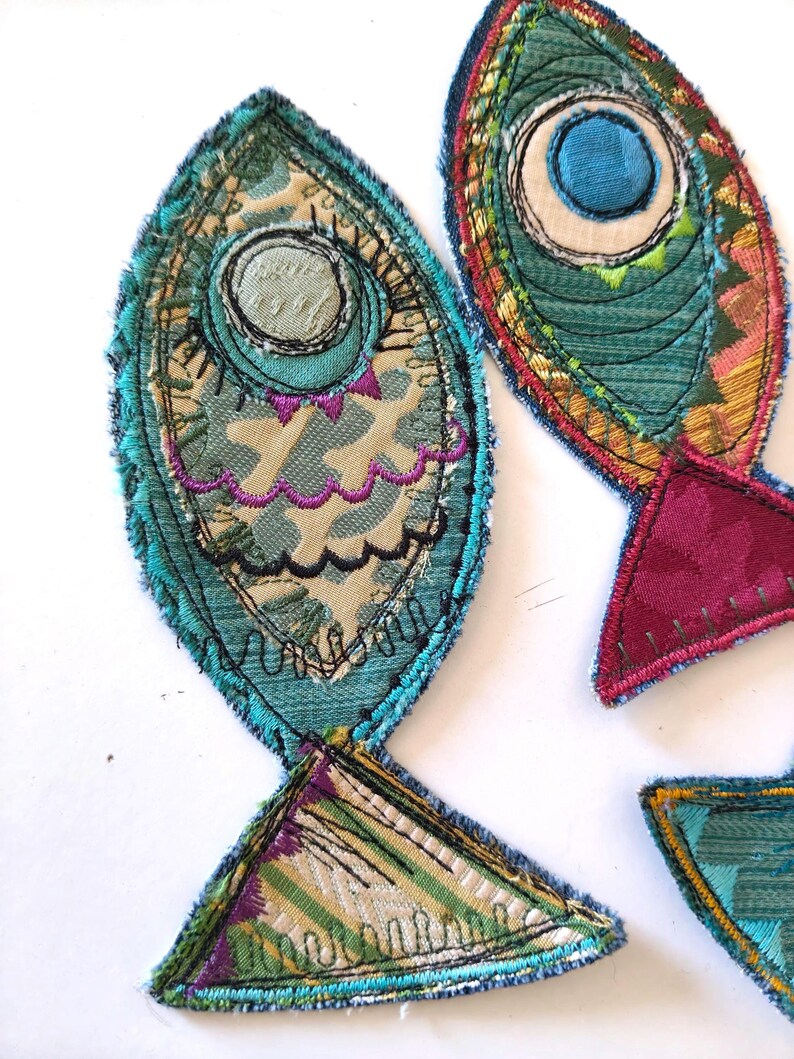 Op de afbeelding: Twee stoffen visapplicaties gemaakt van gelaagde textielresten in tinten blauwgroen, magenta en cr&egrave;me. De visvormen zijn omlijnd met donkerblauw stiksel en bevatten cirkelvormige oogdetails, decoratief schulprandborduurwerk en rafelige randen.