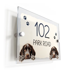 May include: A white and cream house number sign with the number 102 and the text "Park Road" in black. Two brown and white dogs are peeking over the top of the sign. There are paw prints in the top left and right corners of the sign.