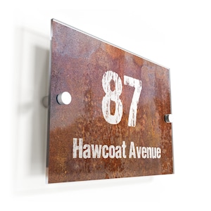 May include: A rusty metal house number sign with white painted numbers and text. The sign reads "87 Hawcoat Avenue".