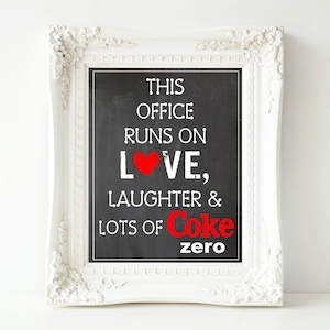 May include: White framed chalkboard sign with the text "This office runs on love, laughter & lots of Coke zero" in black and red lettering. A red heart is in the word "love".
