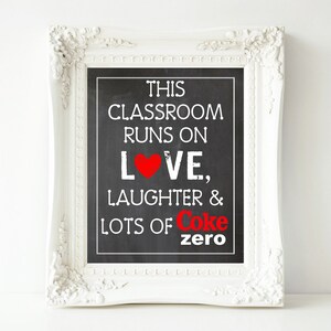 May include: White framed chalkboard sign with the text "This classroom runs on love, laughter & lots of Coke zero" in white lettering with a red heart and the word "Coke" in red.