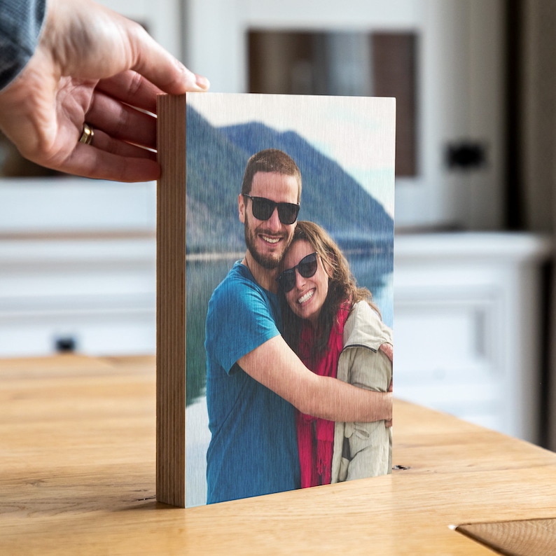 Pu&ograve; includere: Un blocco fotografico in legno con un'immagine stampata di una coppia che sorride e si abbraccia. La coppia indossa occhiali da sole e si trova di fronte a un lago e alle montagne.