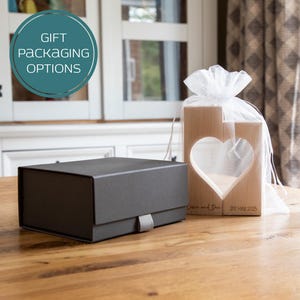 Puede incluir: Una caja de regalo gris oscuro con una cinta gris y un par de bloques de madera con un recorte de coraz&oacute;n. Los bloques est&aacute;n envueltos en una bolsa de malla blanca. El texto "Gift Packaging Options" est&aacute; en un c&iacute;rculo turquesa.