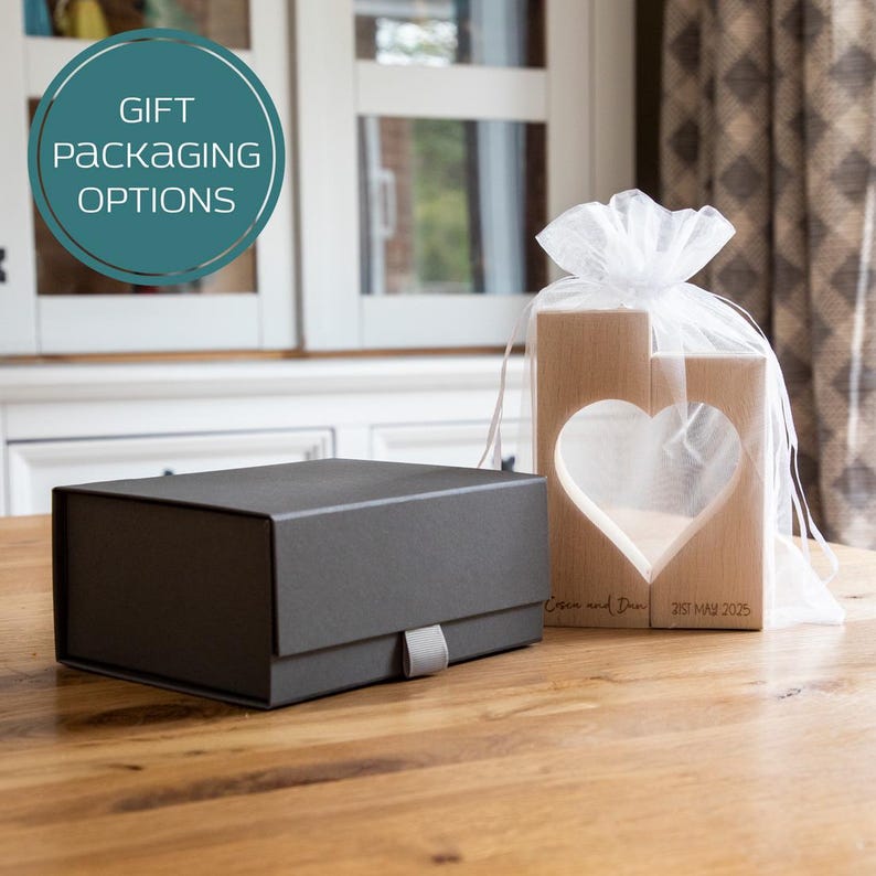 Puede incluir: Una caja de regalo gris oscuro con una cinta gris y un par de bloques de madera con un recorte de coraz&oacute;n. Los bloques est&aacute;n envueltos en una bolsa de malla blanca. El texto "Gift Packaging Options" est&aacute; en un c&iacute;rculo turquesa.
