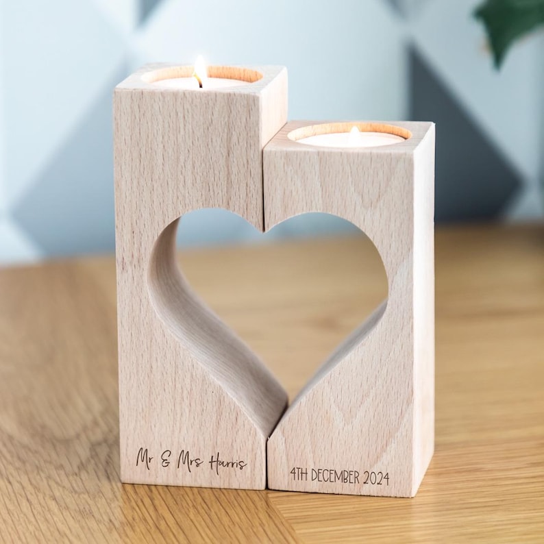 Puede incluir: Un portavelas de madera con dos secciones cuadradas, cada una con una vela encendida. Las secciones est&aacute;n unidas por un recorte en forma de coraz&oacute;n. El texto "Mr & Mrs Harris" y "4TH DECEMBER 2024" est&aacute; grabado en la base.