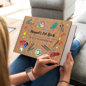 May include: A wooden art book with a spiral binding. The cover features a colourful illustration of art supplies and the text "Abigail's Art Book" and "For all of my experimental work".