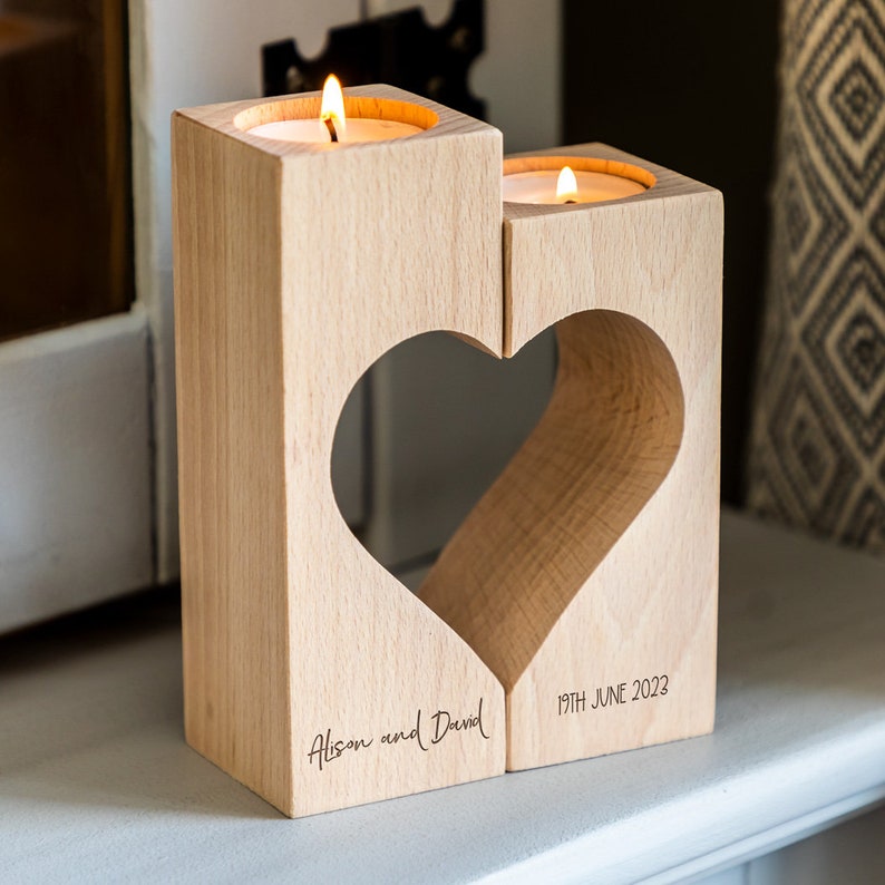 Puede incluir: Un portavelas de madera en forma de coraz&oacute;n con dos velas encendidas. El soporte de color madera natural tiene un recorte en forma de coraz&oacute;n. Los nombres "Alison and David" y la fecha "19 de junio de 2023" est&aacute;n grabados. Un art&iacute;culo decorativo.