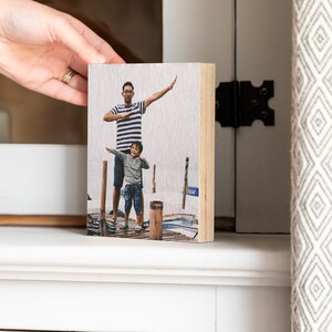 Pu&ograve; includere: Un blocco fotografico in legno con un'immagine stampata di un padre e un figlio in piedi su un molo. Il padre indossa una camicia a righe e il figlio indossa una camicia blu. Il blocco fotografico &egrave; un ottimo modo per esporre una foto preferita.