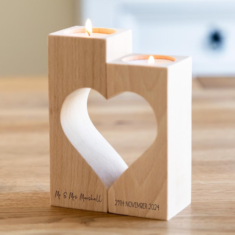Puede incluir: Portavelas de madera con dos velas encendidas. Una forma de coraz&oacute;n est&aacute; recortada en la madera. El texto "Mr & Mrs Marshall" y "29TH NOVEMBER 2024" est&aacute;n impresos en la base del portavelas.