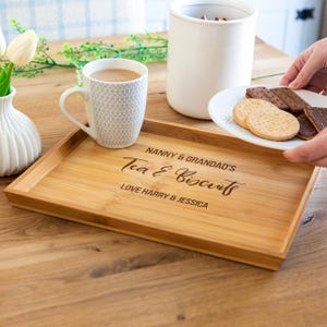 May include: A rectangular bamboo serving tray engraved with "Nanny & Grandad's Tea & Biscuits Love Harry & Jessica". A mug of tea, a white vase with flowers, and a white container are on the tray. A plate of biscuits is held nearby.