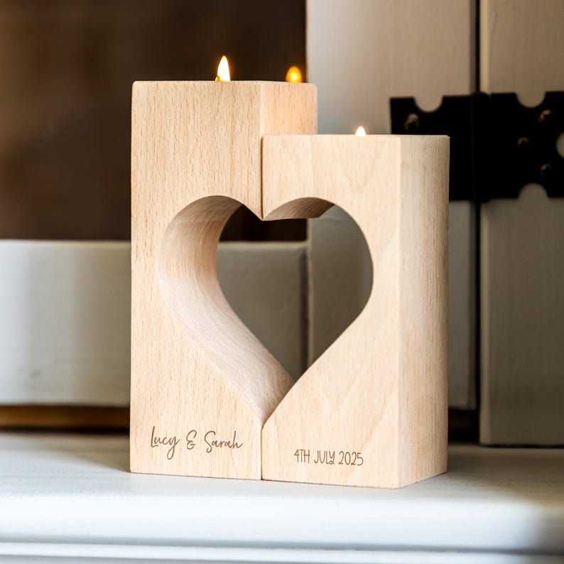 Puede incluir: Un portavelas de madera con forma de coraz&oacute;n, compuesto por dos piezas. Cada una sostiene una vela encendida. Los nombres "Lucy & Sarah" y la fecha "4th July 2025" est&aacute;n grabados en la madera.