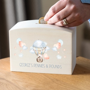 May include: A light beige wooden money box with a whimsical design featuring a hot air balloon with a small animal inside, surrounded by pink and blue hearts and stars. The text "GEORGE'S PENNIES & POUNDS" is printed on the front.