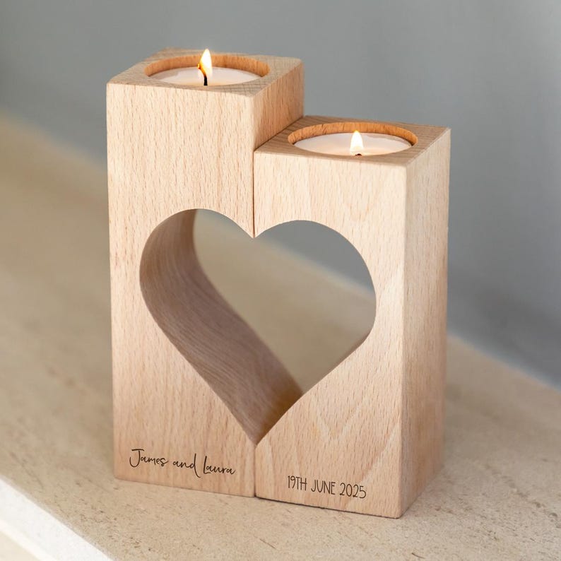 Puede incluir: Un portavelas de madera clara con dos espacios para velas y un recorte en forma de coraz&oacute;n. Las velas est&aacute;n encendidas. El portavelas est&aacute; grabado con los nombres "James and Laura" y la fecha "19TH JUNE 2025".