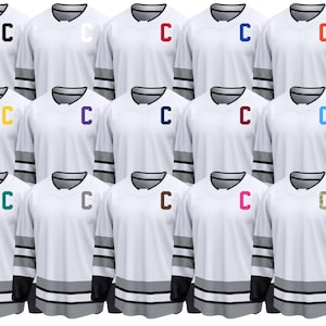 Peut inclure: Plusieurs maillots de hockey blancs à manches longues, chacun avec un logo "C" de couleur différente sur la poitrine. Les maillots ont des manches rayées grises et noires et un col en V. Les logos "C" sont de couleurs variées.