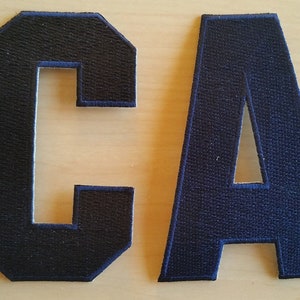 May include: Two felt fabric letters, "C" and "A", in navy blue with a white outline. The letters are sewn together and are approximately 4 inches tall.
