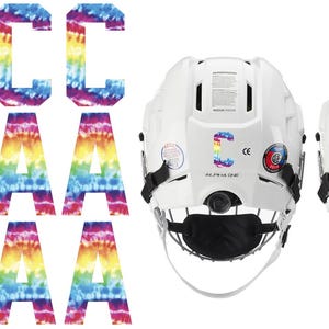 May include: White hockey helmets with tie-dye letter decals. The letters "C" and "A" are displayed on the helmets. The tie-dye letters are in rainbow colors. Additional tie-dye letters are also shown.